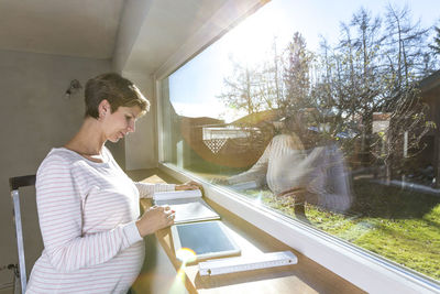 Pregnant woman reading document by window at home on sunny day