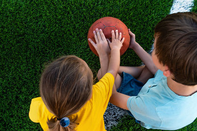 Unrecognizable children sitting on green grass and holding ball outdoors