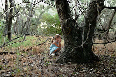 Side view of girl crouching by tree trunk in forest