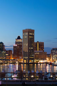 Sailboats moored in harbor by modern illuminated buildings against clear sky at dusk