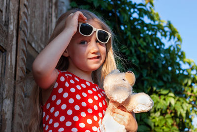 Beautiful happy girl in red polka dot dress soft toy on wooden balcony. cute joyful child long hair.