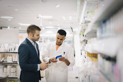 Male pharmacist showing medicine to mature customer standing by rack at medical store