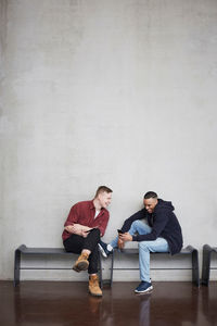 Full length of happy male students with technologies sitting on bench against wall in university