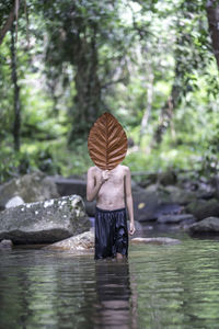 Shirtless man holding leaf while standing in lake against trees