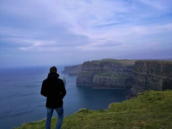 Rear view of man standing on cliff by sea against sky