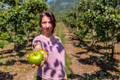 Portrait of smiling woman holding apple against trees