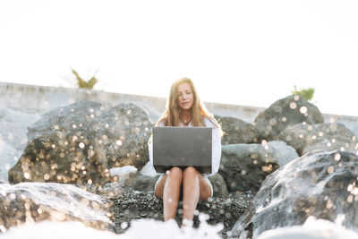 Young freelance businesswoman with long hair in a white shirt working at laptop on seashore