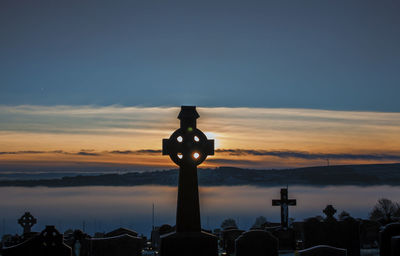 Cross and tombstones in graveyard against sky during sunset