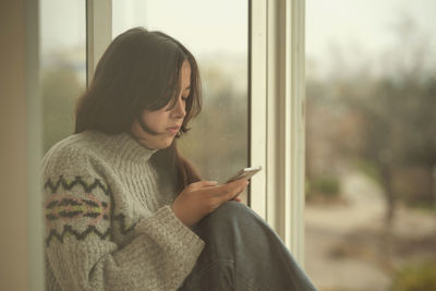 Young woman using mobile phone at window