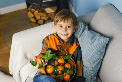 Boy peels and eats a tangerine, sitting in an armchair by the fireplace.