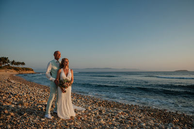 Bride and groom standing on beach against clear sky