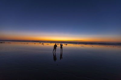 Silhouette people at beach against dramatic sky during sunset