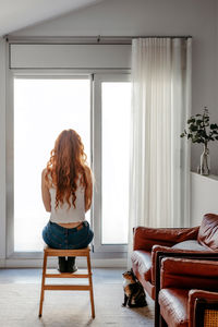 Back view full body of unrecognizable female with red hair sitting alone against panoramic windows in light living room with sofas