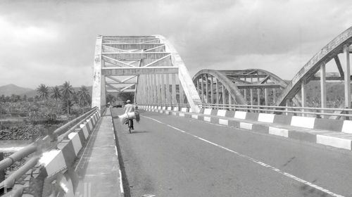 Rear view of man riding bicycle on bridge against cloudy sky