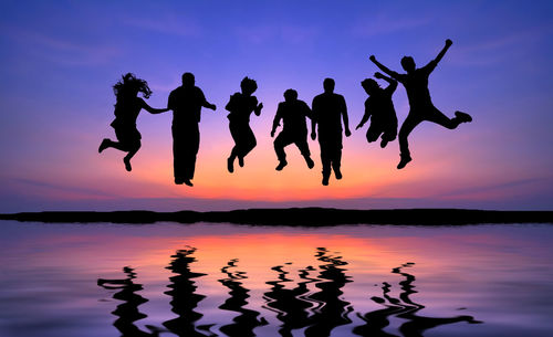 Silhouette people jumping by lake against sky during sunset