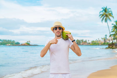 Portrait of male tourist pointing at coconut by seashore