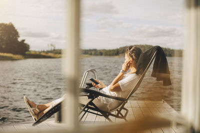 Full length of woman wearing headphones while using mobile phone on patio seen through window