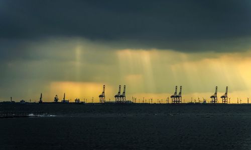 Silhouette factory by sea against sky during sunset