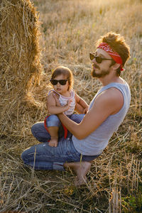 Father and son in t-shirts sitting next to a haystack on a sloping field during sunset