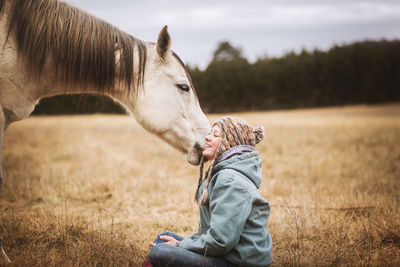 White horse tugging on girls hat in field in the fall while girl sits