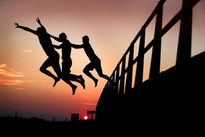 Silhouette men jumping from railing against sky during sunset