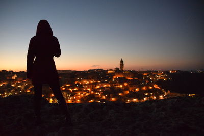 Rear view of silhouette man standing by illuminated buildings against sky during sunset