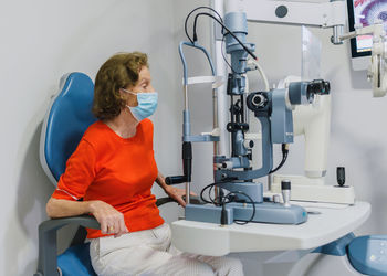 Side view of senior female patient in protective medical mask sitting at ophthalmological equipment of ophthalmoscope