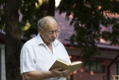 An elderly man with glasses reads a book in the park in the fresh air.