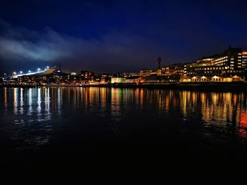 Illuminated buildings by river against sky at night