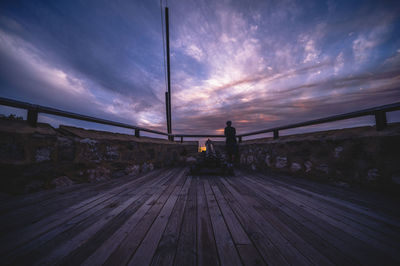 Rear view of man standing on railing against sky during sunset