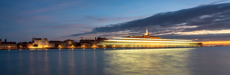 Blurred motion of illuminated cruise ship on sea by city at dusk