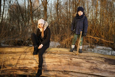 Full length portrait of mother and son on fallen tree during winter