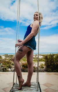 Full length portrait of young woman standing on swing against sky