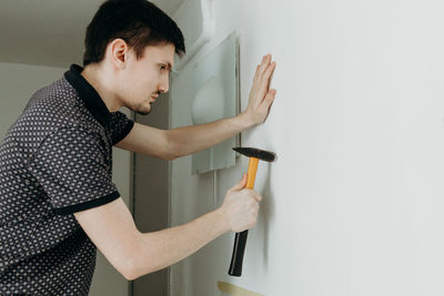 A young man hammers a dowel into a hole on the wall.