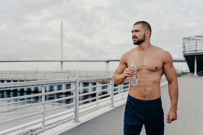 Shirtless man holding water bottle while standing on bridge