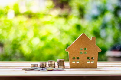 Close-up of cardboard model home with coins on wooden table against plants