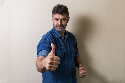 Portrait of mature man gesturing thumbs up while standing against wall