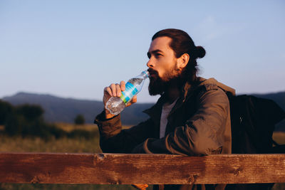 Young man drinking water from bottle while sitting on wood against sky