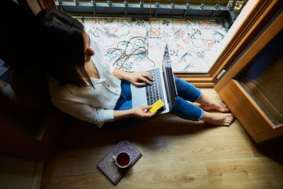 Top view of young woman shopping and paying online on a laptop with credit card while relaxing at home next to a window.