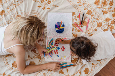 High view of caucasian mom and hispanic daughter painting while they are lying in the bed.