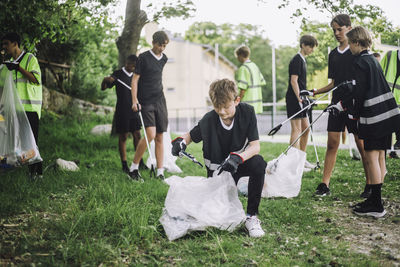 Boy kneeling while collecting garbage in plastic bag near friends