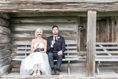 Portrait of bride and groom holding artificial mustache and lips while sitting on bench