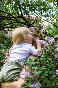 Portrait of young caucasian mother who is holding her son and touching flowers of lilac tree. 