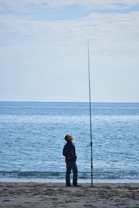 Full length of man looking at fishing rod at beach against sky