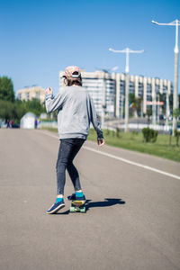 Rear view of man skateboarding on road against clear blue sky during sunny day