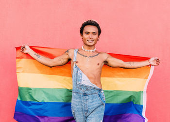 Portrait of smiling man holding rainbow flag standing in front of pink wall
