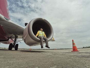 Full length of worker sitting on airplane jet engine at runway