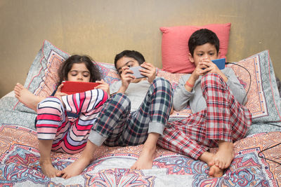 Full length of siblings using mobile phones on bed