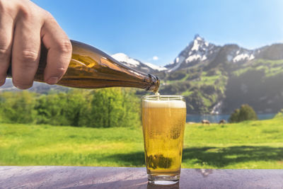 Cropped hand pouring beer from bottle in glass against mountain
