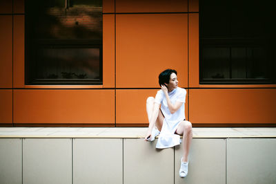 Full length of young woman sitting on walkway against orange building